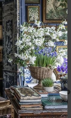 A table with a large floral arrangement in a wicker basket, featuring white flowers, purple irises, and green foliage, surrounded by books and decorative items, with framed artwork and a mirror on the wall in the background.