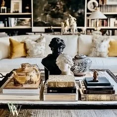 Decorative objects including bust sculptures, vases, and ornate boxes arranged on a table in front of a sofa with yellow and white pillows, in a modern living room with large windows.