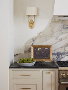 Kitchen countertop with a framed painting, a white bowl of green decorative objects, small cabinet drawers, and a marble backsplash.