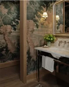 Bathroom sink with a mirror, wall sconces, and a vase of green flowers, in front of a nature-themed wallpaper.