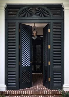 Black double doors with a fan-shaped transom window and louvered shutters, opening to a decorated interior with a hanging lantern light.