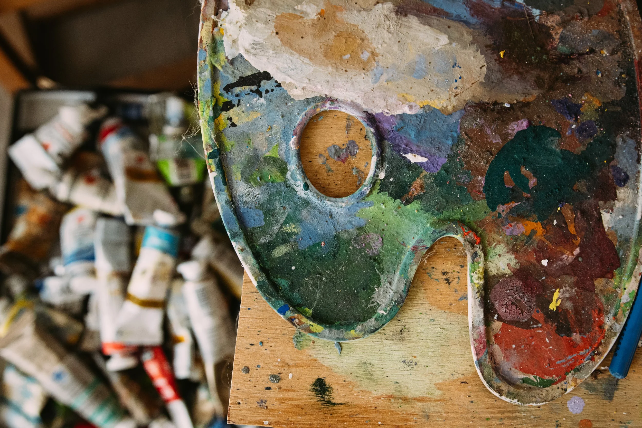 A worn artist's palette with colorful paint stains and two circular cutouts, placed on a wooden surface, with scattered paint tubes in the background.