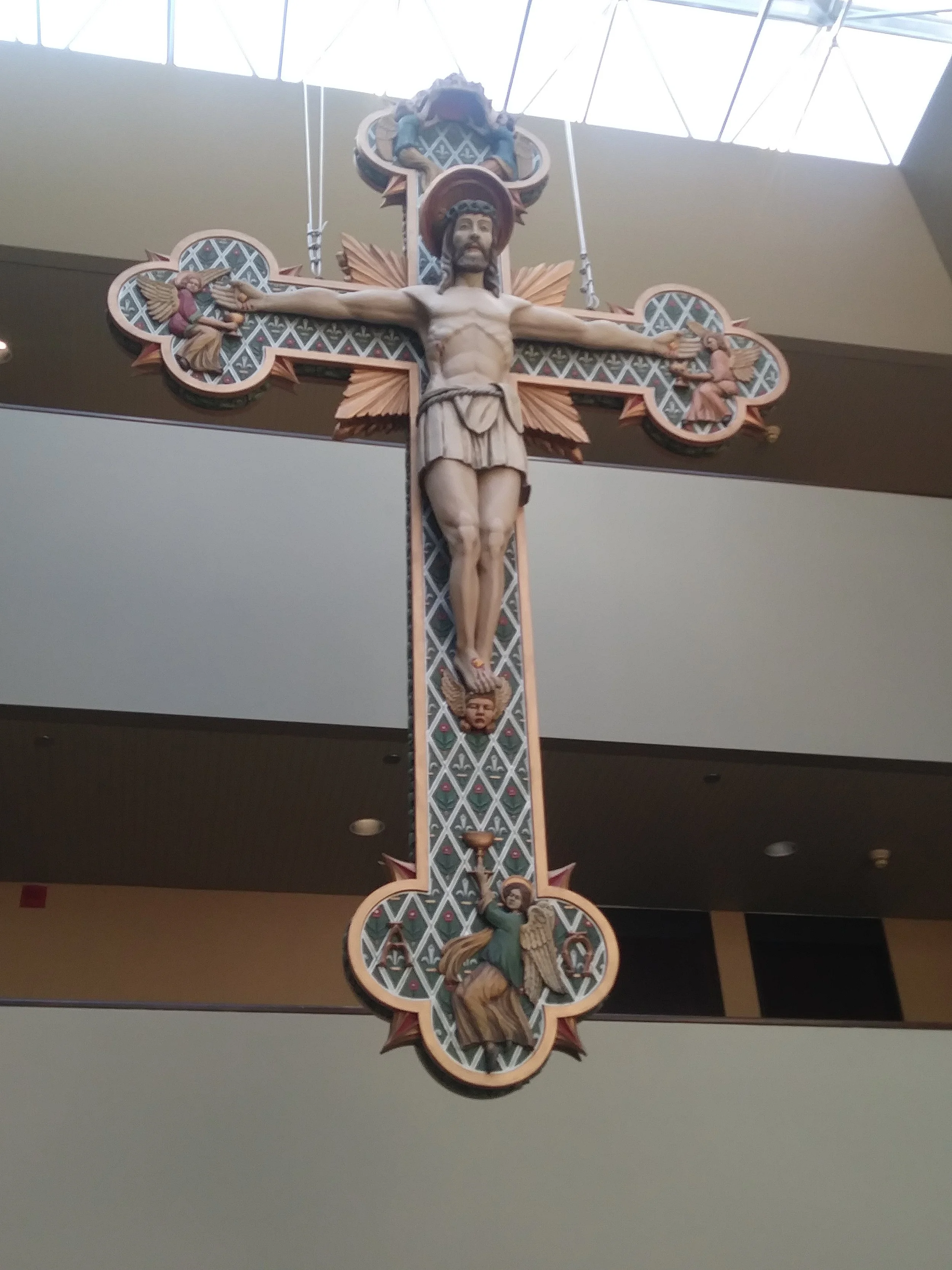 Decorative crucifix with carved figure of Jesus Christ on a cross, featuring angels and intricate design details, hanging inside a building with a glass ceiling.