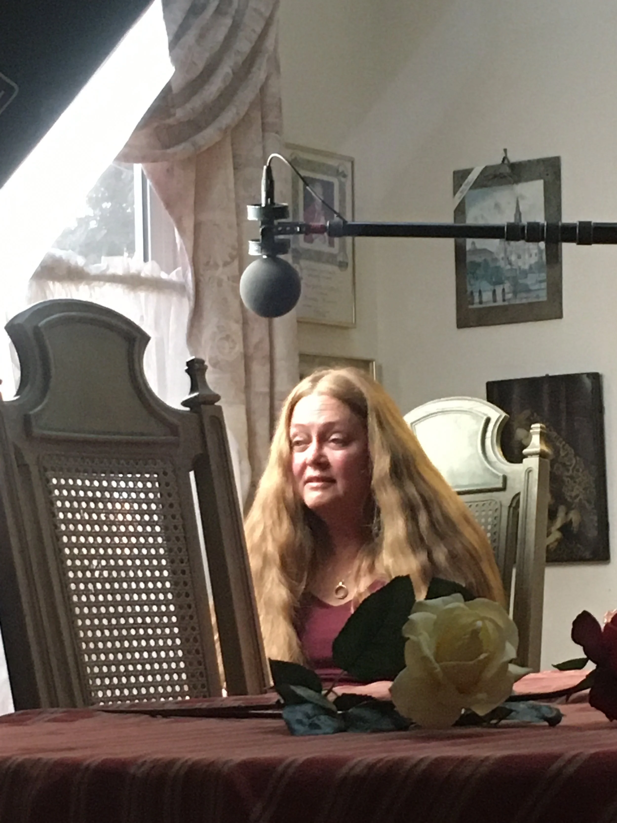 A woman with long, blonde hair sitting at a table with a tablet, a large white rose, black roses, and some papers. In the background, there are paintings on the wall and a window with a sheer curtain.