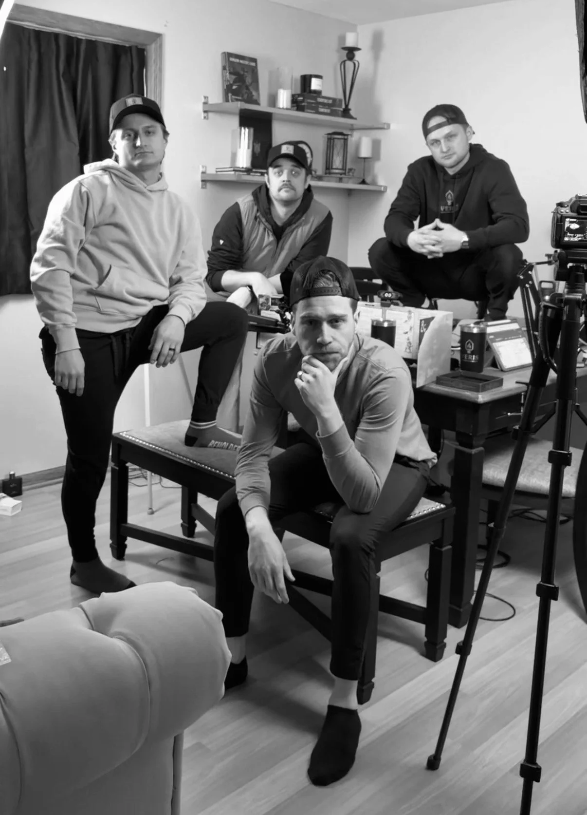 Black and white photo of four young men in a room with photography equipment, furniture, and shelves, posing for a photo.