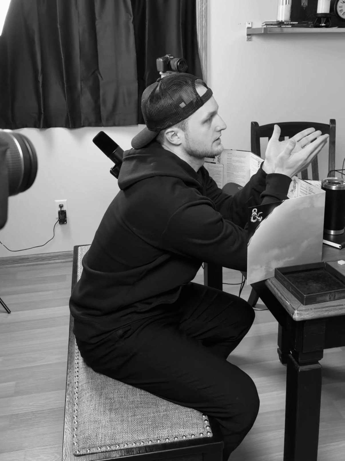 A man wearing a baseball cap and a hoodie is sitting at a table, in a room with a camera and a microphone set up nearby, appearing to be in conversation or explanation.