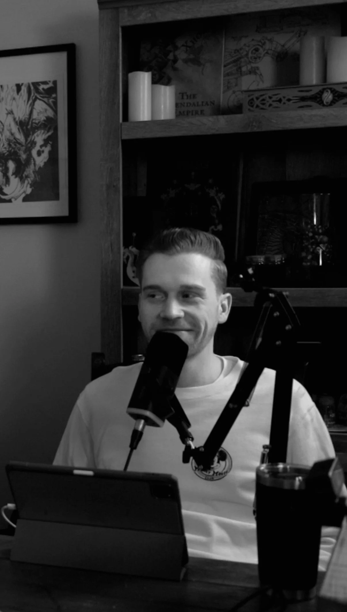 A man with short, styled hair sits in front of a microphone, smiling slightly, in a room with bookshelves and framed pictures.