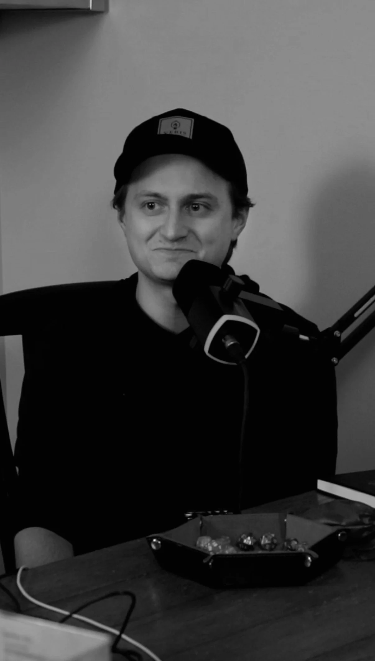 A man sitting at a desk, wearing a black cap, speaking into a microphone, with a bowl of chocolates in front of him, in a monochrome setting.