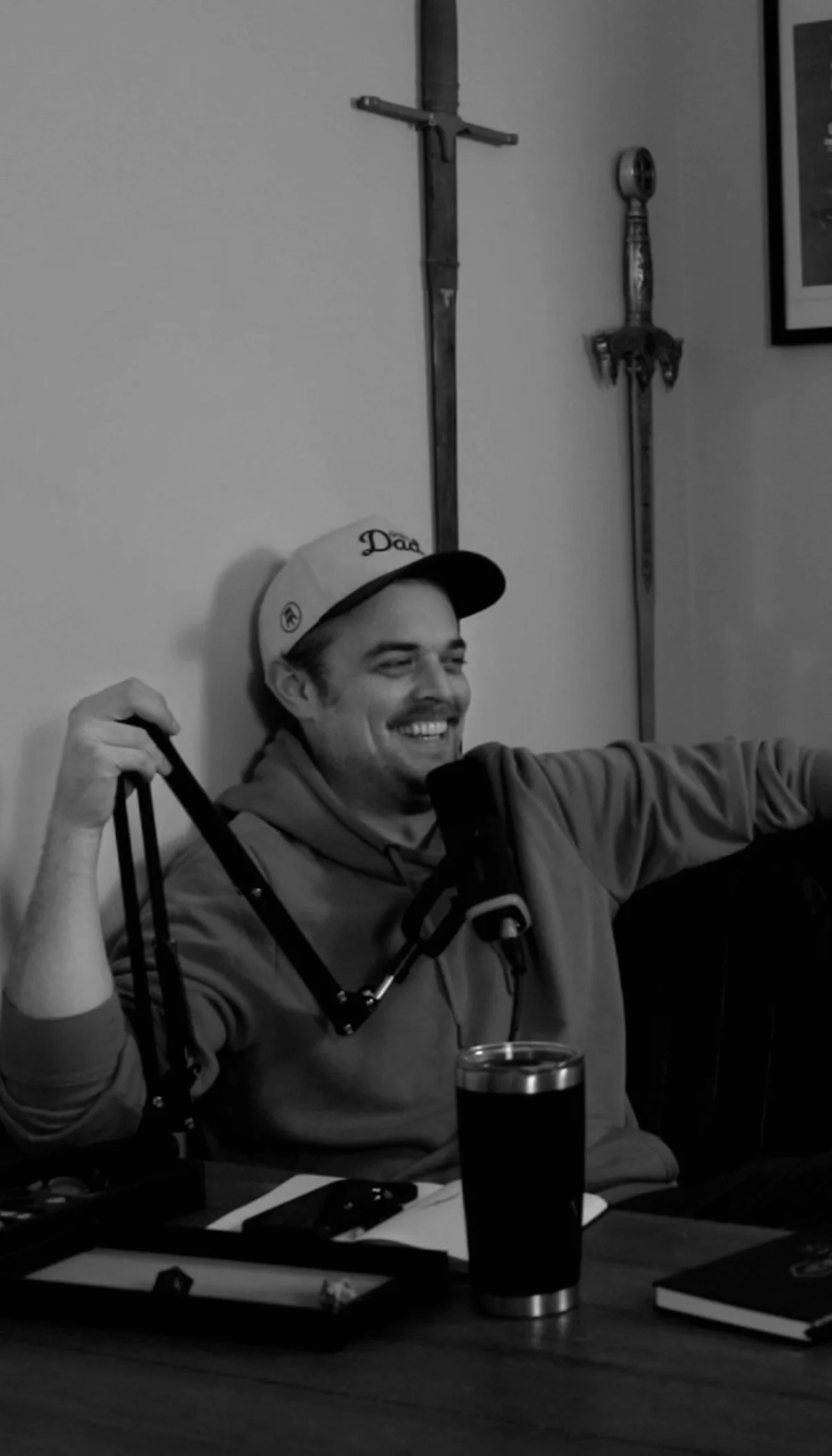 A young man sitting at a table, smiling, wearing a baseball cap and hoodie. The table has a glass filled with dark liquid, some books, and other items. Swords are mounted on the wall behind him.