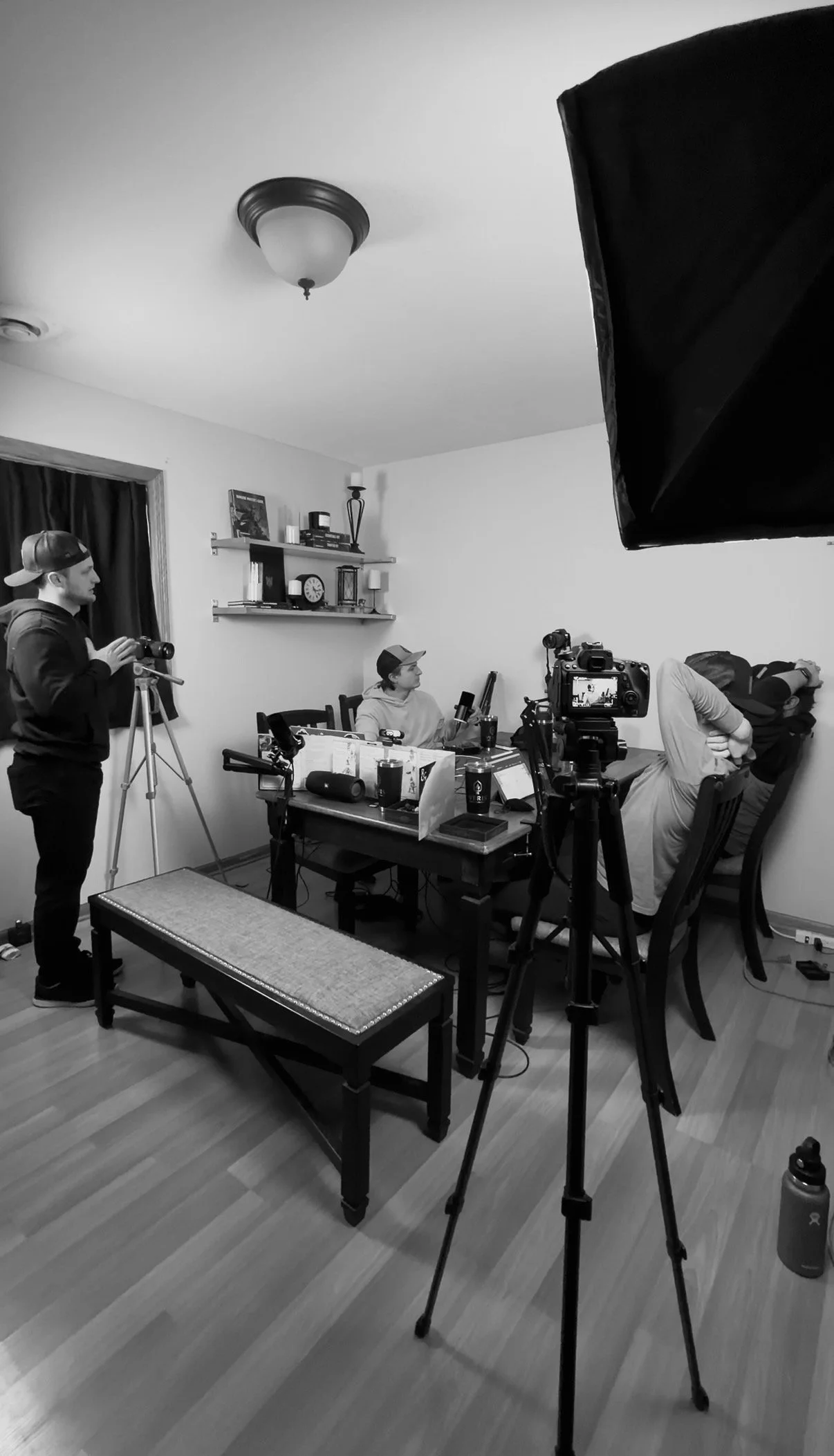 A black and white photo of a home recording or streaming setup with three people; two sitting at a table with cameras, one standing adjusting equipment; various cameras, microphones, and a computer on the table; shelves with books and decor on the wall; a water bottle on the floor.