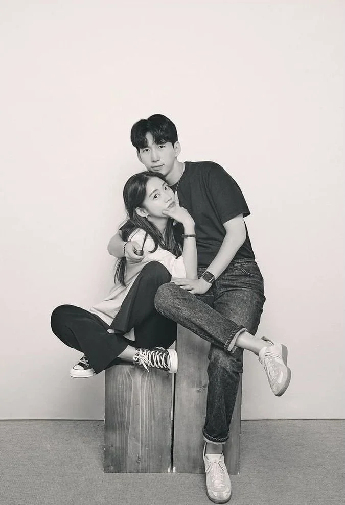 Two young people, a man and a woman, sitting on wooden blocks against a plain wall. The woman is sitting cross-legged, wearing sneakers and casual clothes, resting her chin on her hand. The man is standing, wearing jeans and sneakers, with one leg crossed over the other and one arm around her shoulders. The photo is in black and white.