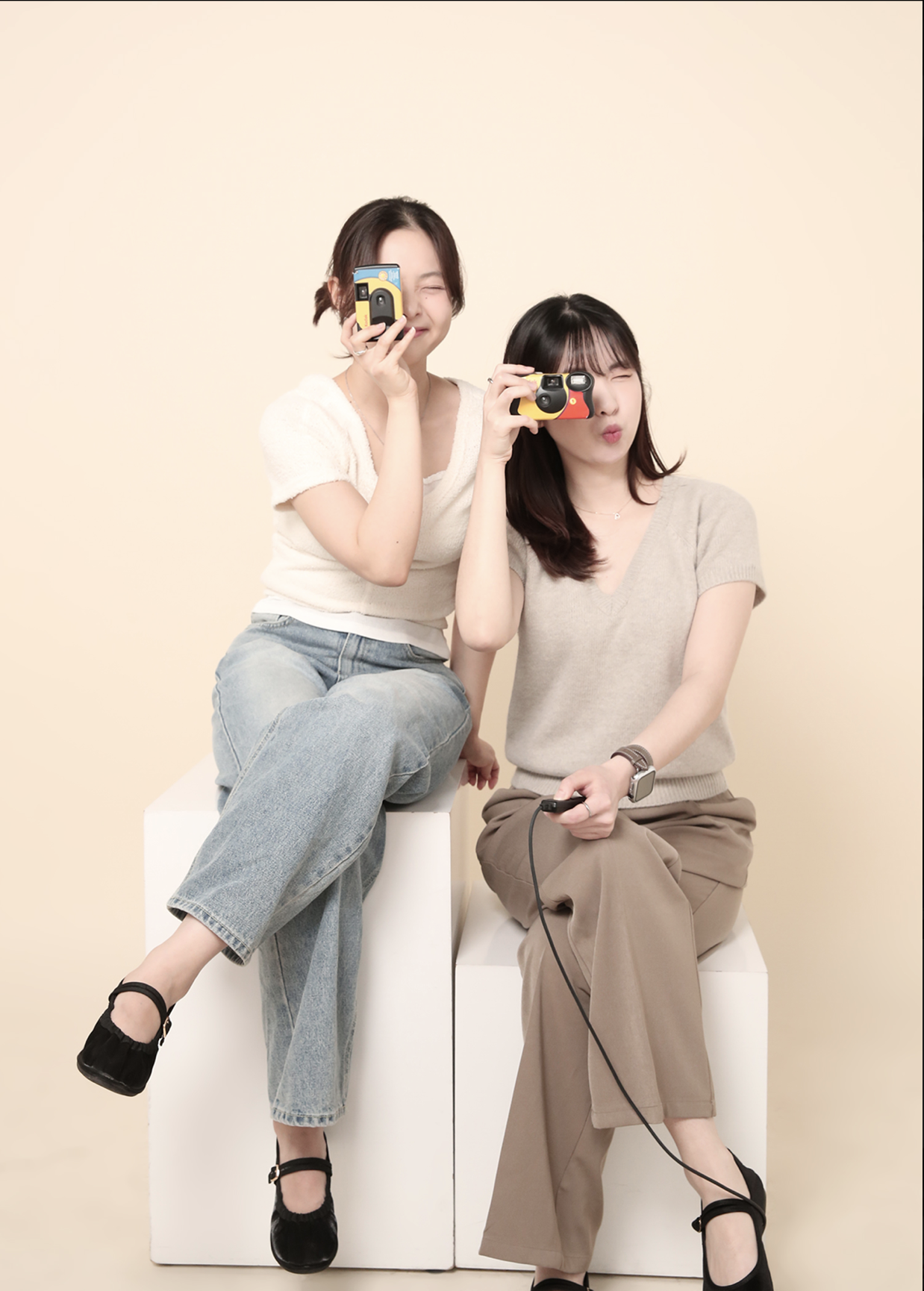 Two women posing with toy cameras against a plain beige background. One woman is sitting on a white block with crossed legs, smiling and covering one eye, while the other woman is seated on a white block, puckering her lips and holding a camera to her eye. Both women are casually dressed.