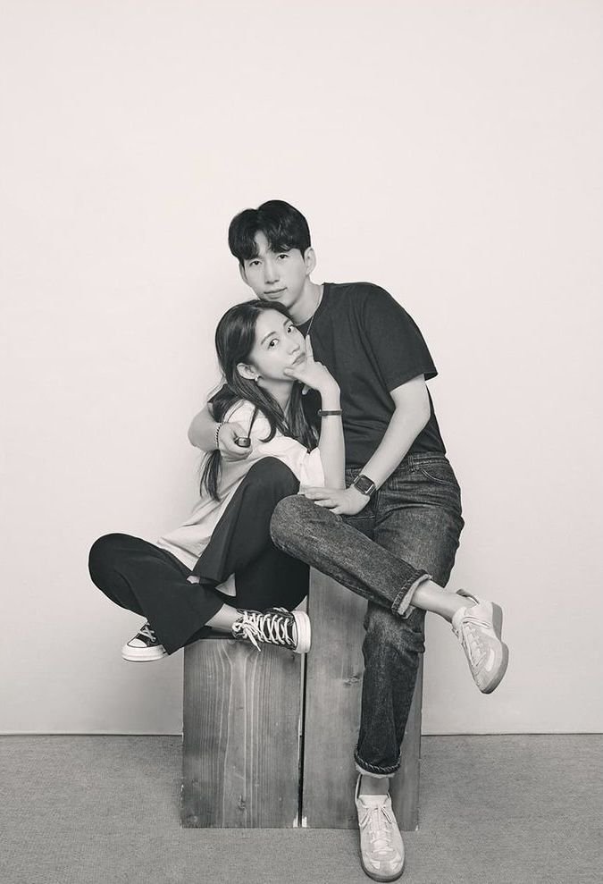 A black and white photo of a young man and woman sitting on wooden blocks, posing together with the woman sitting cross-legged and the man standing behind her, both with relaxed expressions.