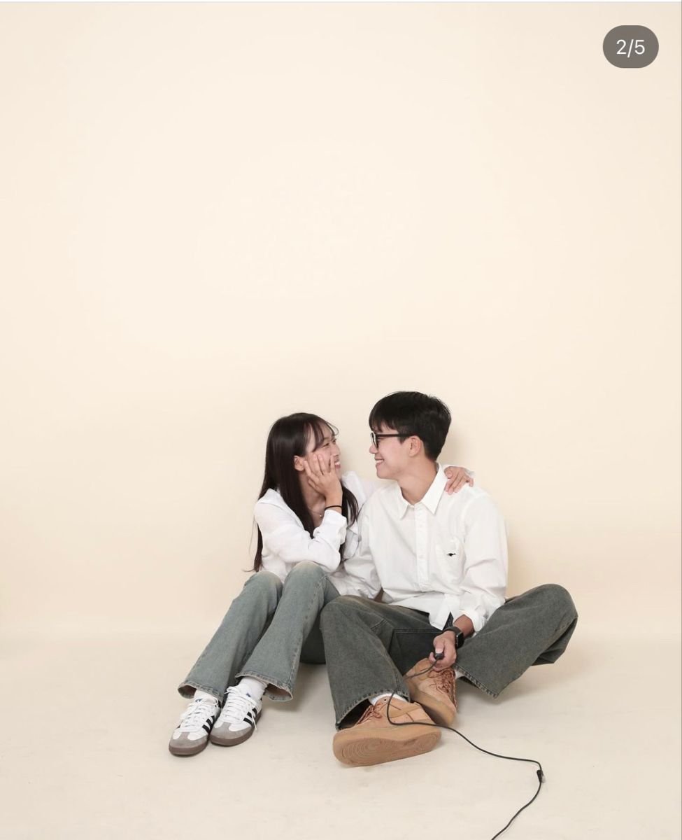 A young man and woman sit on the floor against a plain light-colored background, smiling and looking at each other. The woman gently touches the man's face, and they appear to be happy and close.