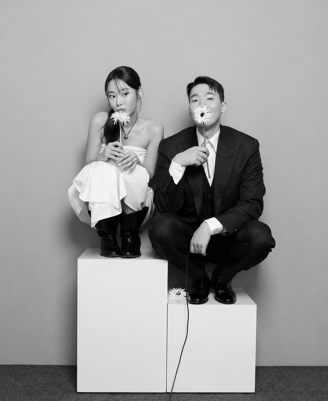 A black-and-white photo of a young man in a suit sitting cross-legged on a podium with a flower in his mouth, and a young woman in a strapless dress and boots sitting on a higher podium with a flower in her mouth, both against a plain background.