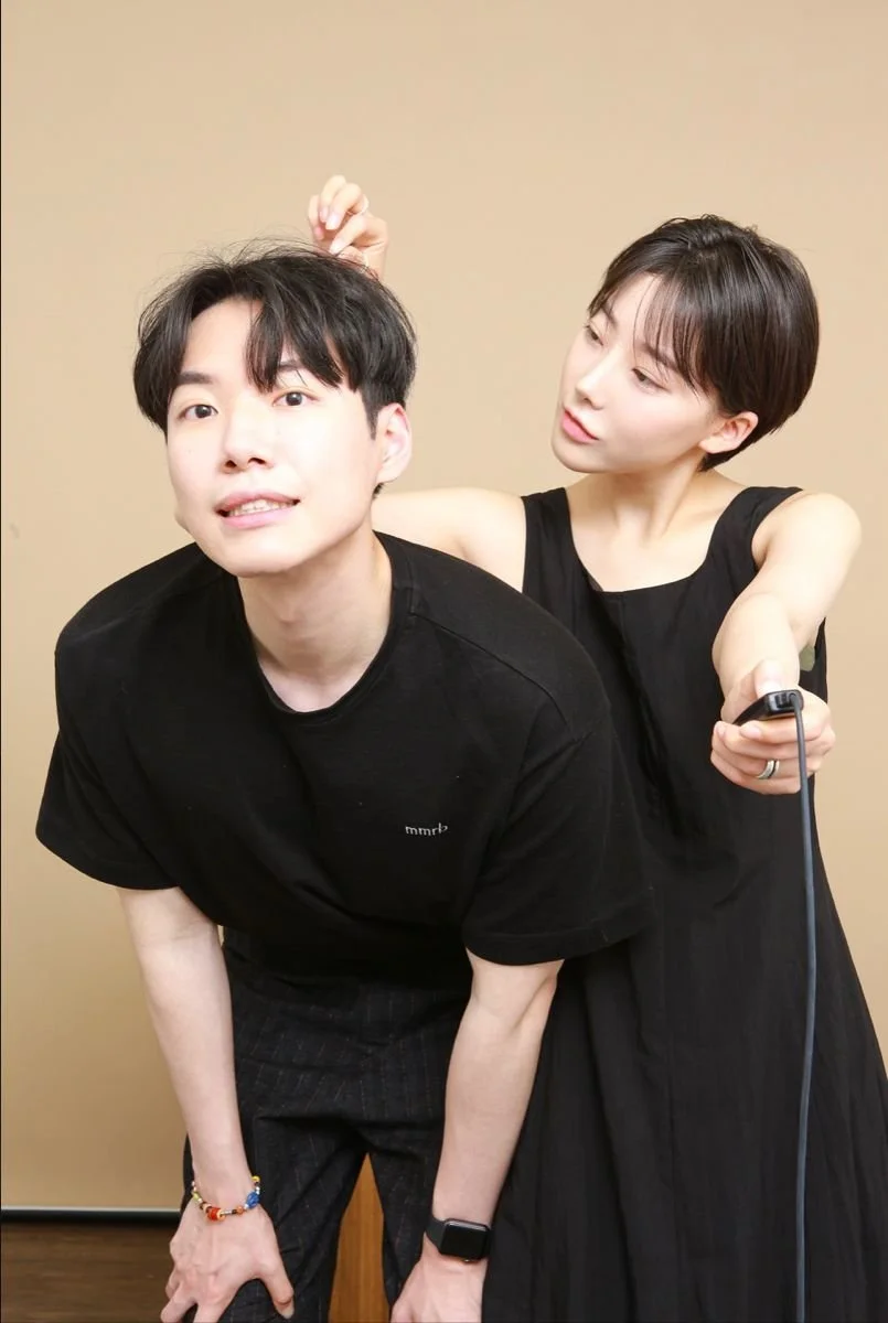 A man and a woman are posing for a photo. The woman is styling the man's hair while holding a straightening iron. They are both wearing black clothing and are in front of a beige background.