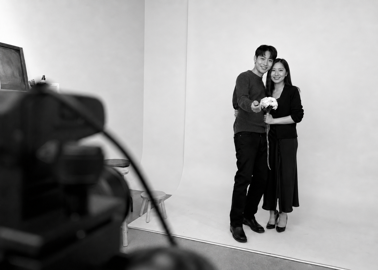 A black-and-white photo of a man and woman standing together in front of a plain background, smiling. The man is wearing a dark sweater and pants, holding a bouquet of flowers, while the woman is wearing a dark top and pants. They are close, with the woman's arm around the man's waist, and the man pointing toward the camera.