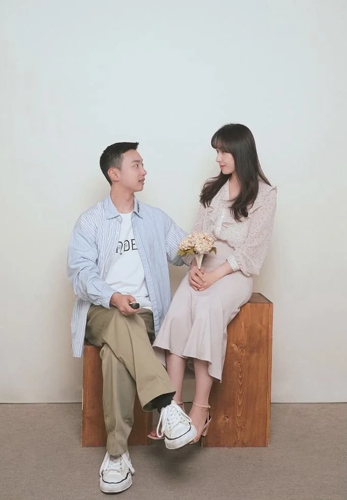 A young man and woman sitting on wooden blocks against a plain wall. The man is holding a remote and looking at the woman, who is holding a bouquet of flowers and looking down at him.