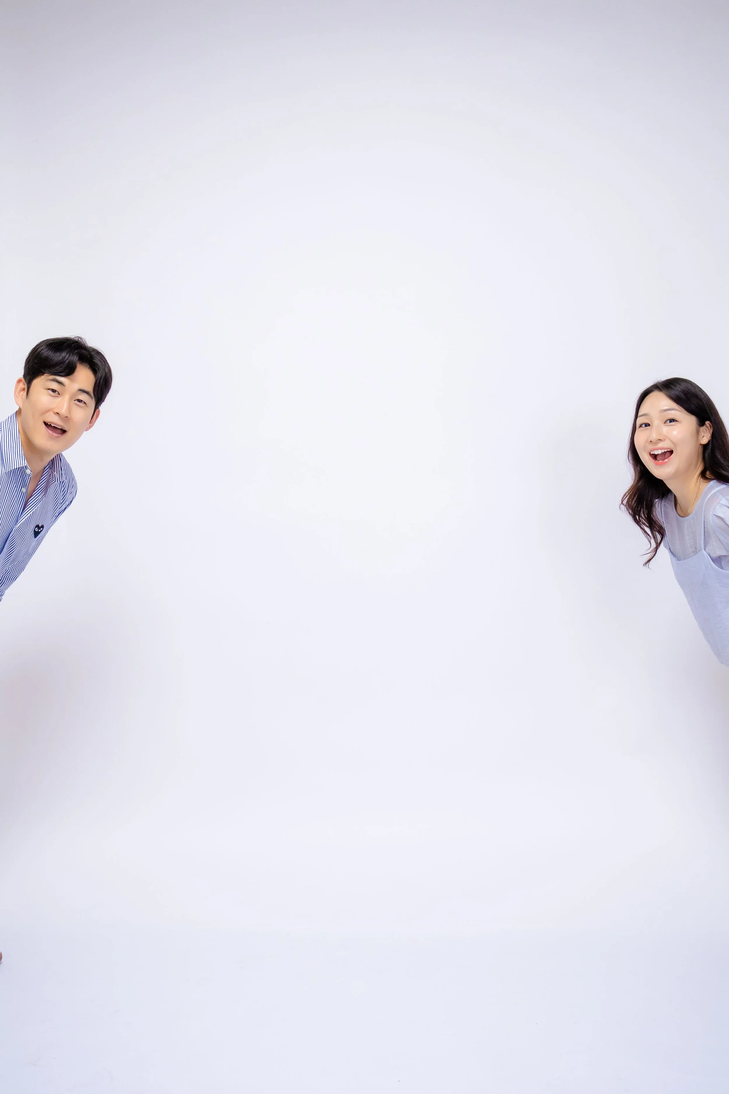 A young man and woman sitting on stools, holding open books above their heads, smiling and facing forward against a light blue background.