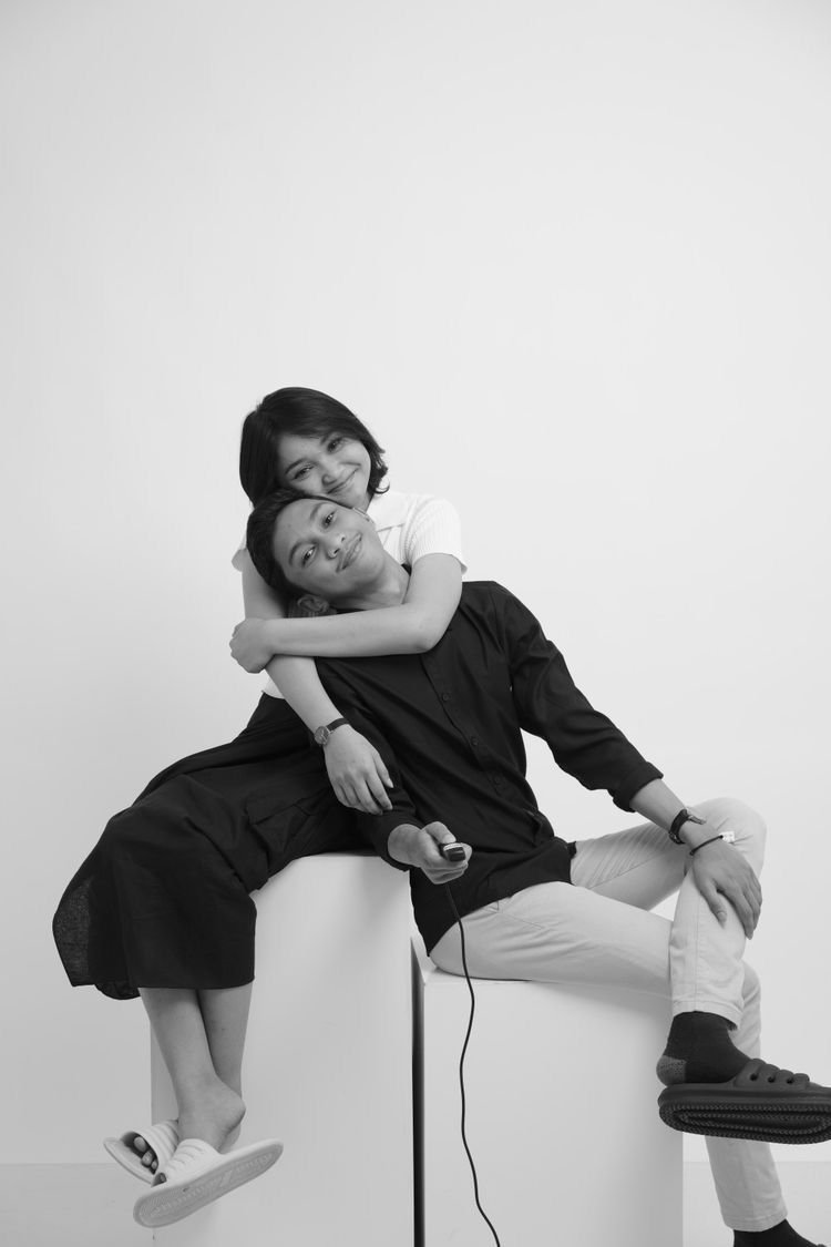 Black and white photo of a young woman hugging a young man who is seated, both smiling, with a plain white background.