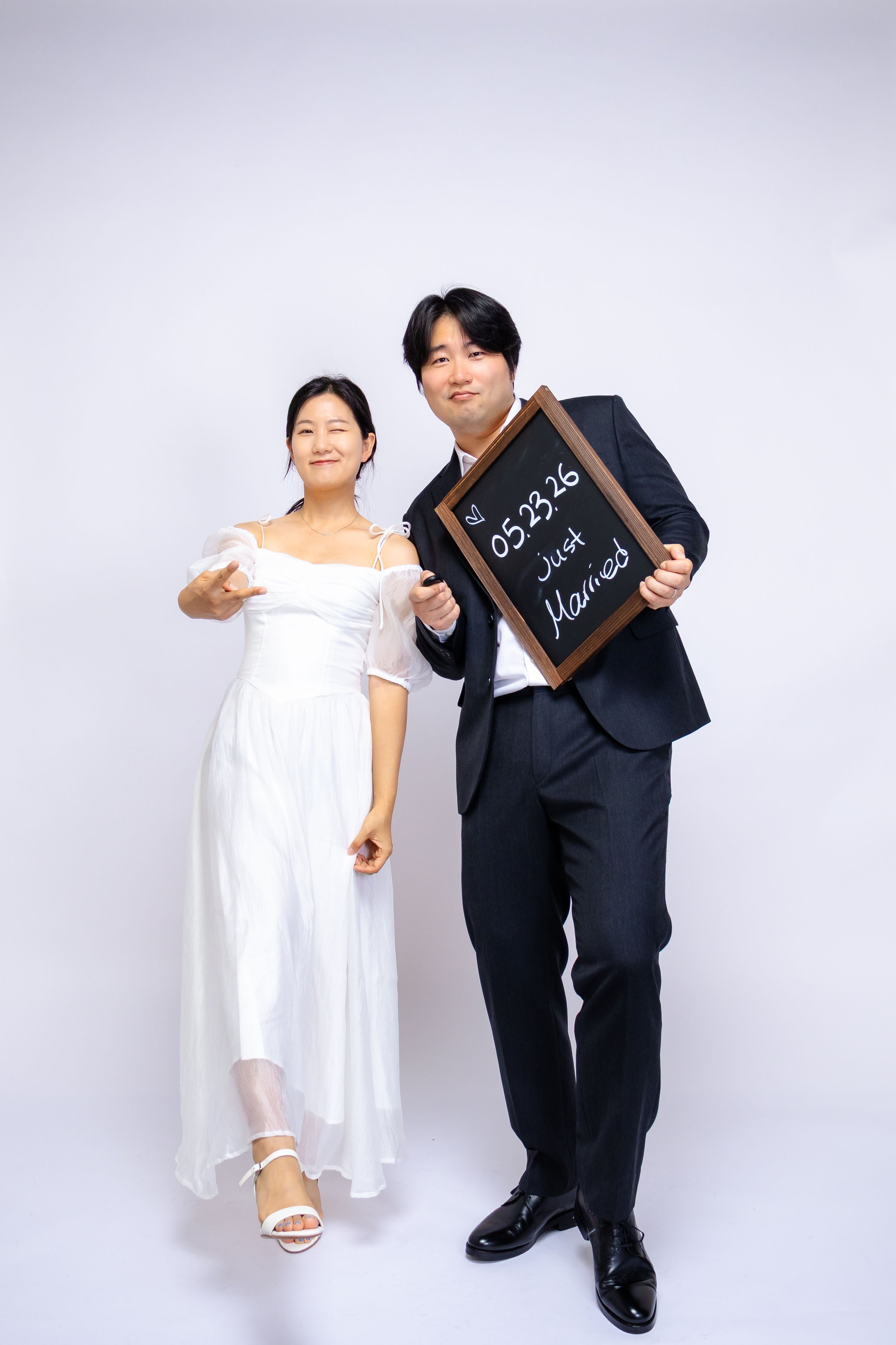 A couple, dressed in wedding attire, standing against a plain white background. The woman is in a white wedding dress with off-shoulder sleeves, winking and smiling. The man is in a dark suit, holding a blackboard that has the date '05.23.26' and the words 'Just Married' written on it, posing with a slight smile.