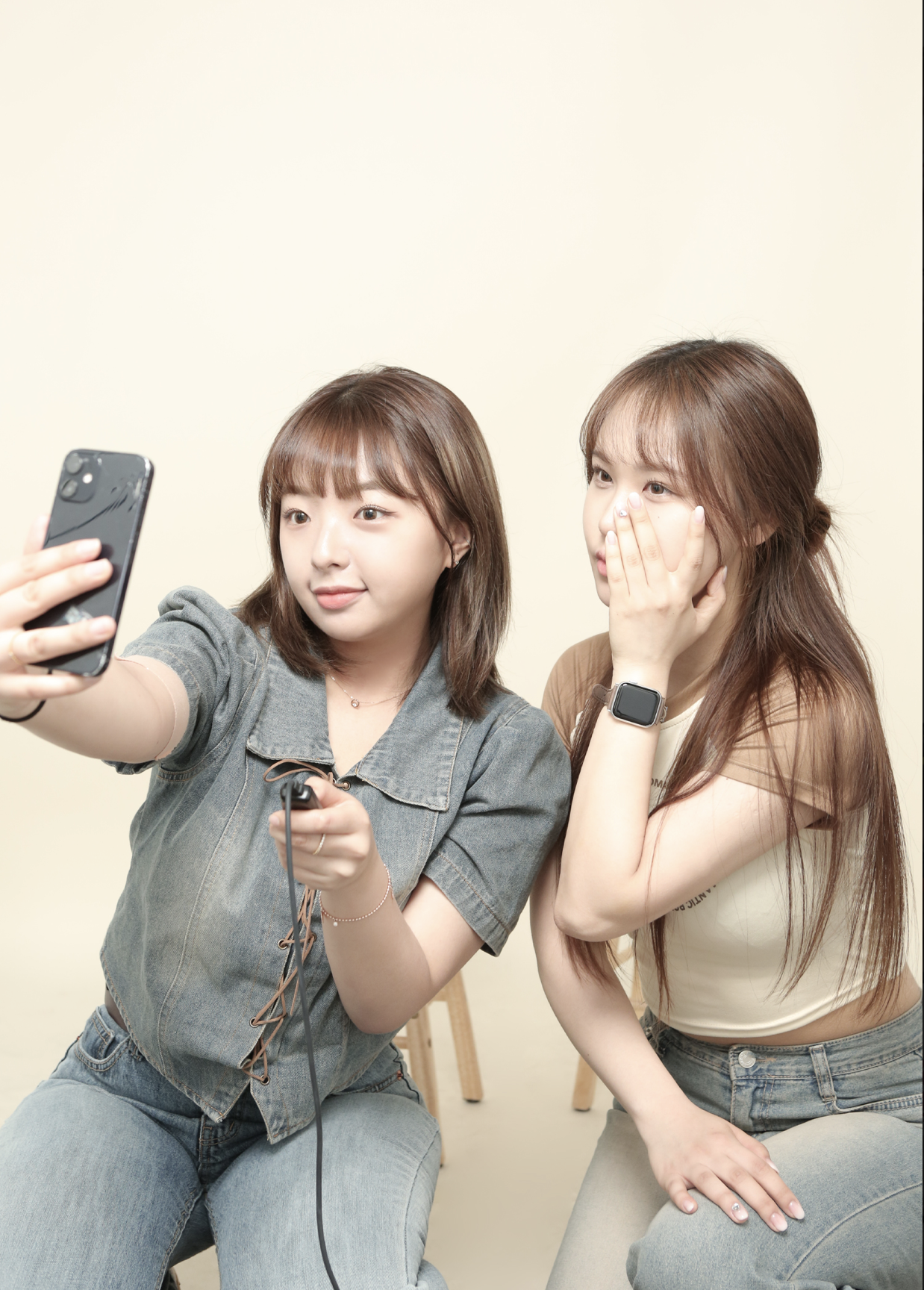 Two women taking a selfie together. One is holding a smartphone and the other is covering her mouth with her hand, both smiling. They are sitting on stools against a plain light background.