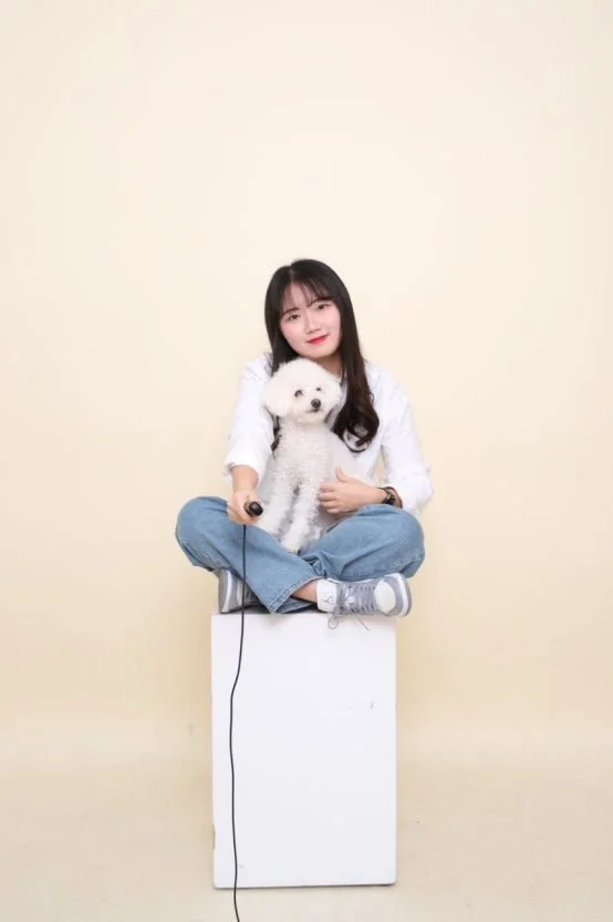 A young woman with long dark hair sitting cross-legged on a white box, holding a white fluffy dog, with a neutral beige background.