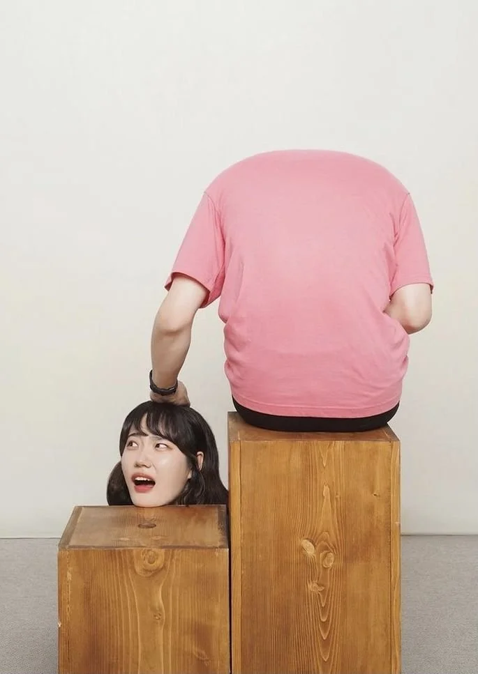 Person with pink shirt sitting on wooden blocks, with a woman's head emerging from a hole in the lower block, her face showing surprise.