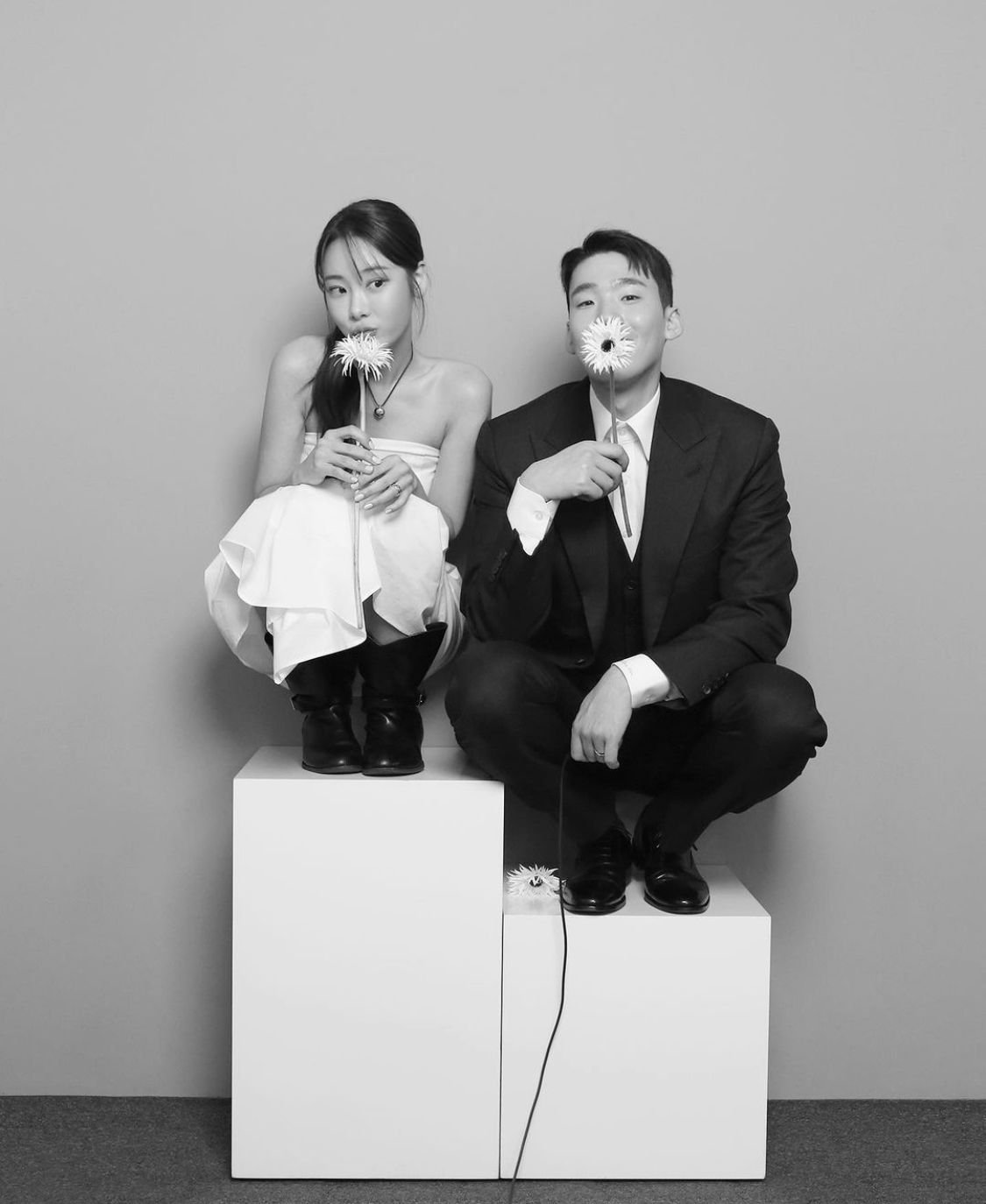 Black and white photo of a woman and a man sitting on white blocks, holding flowers in front of their faces, posed against a plain wall.