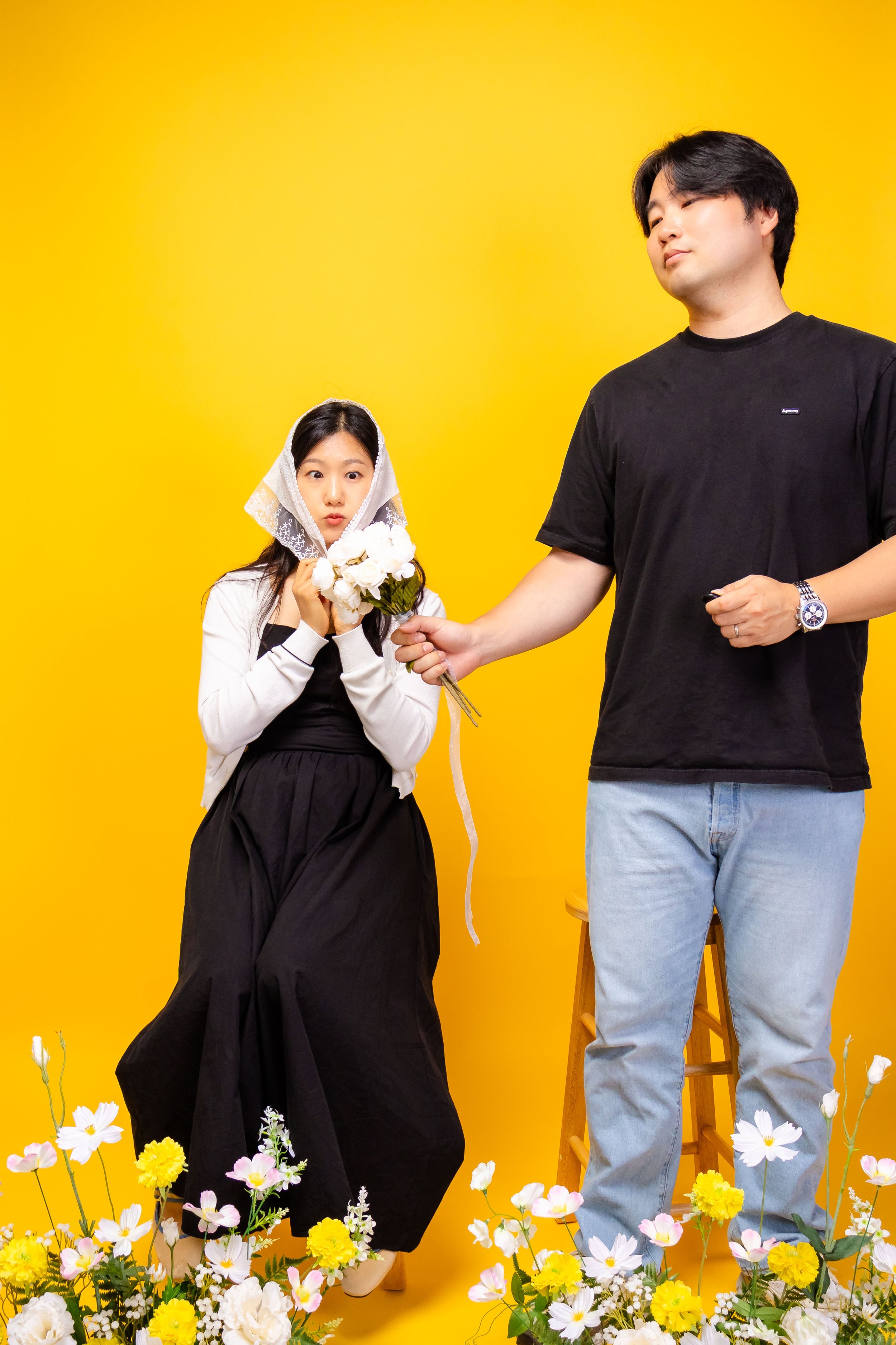 A young woman holding a bouquet of white flowers with a white lace head covering, standing in front of a yellow background, and a young man in a black T-shirt and light blue jeans offering her a flower. There are colorful flowers at the bottom of the image.