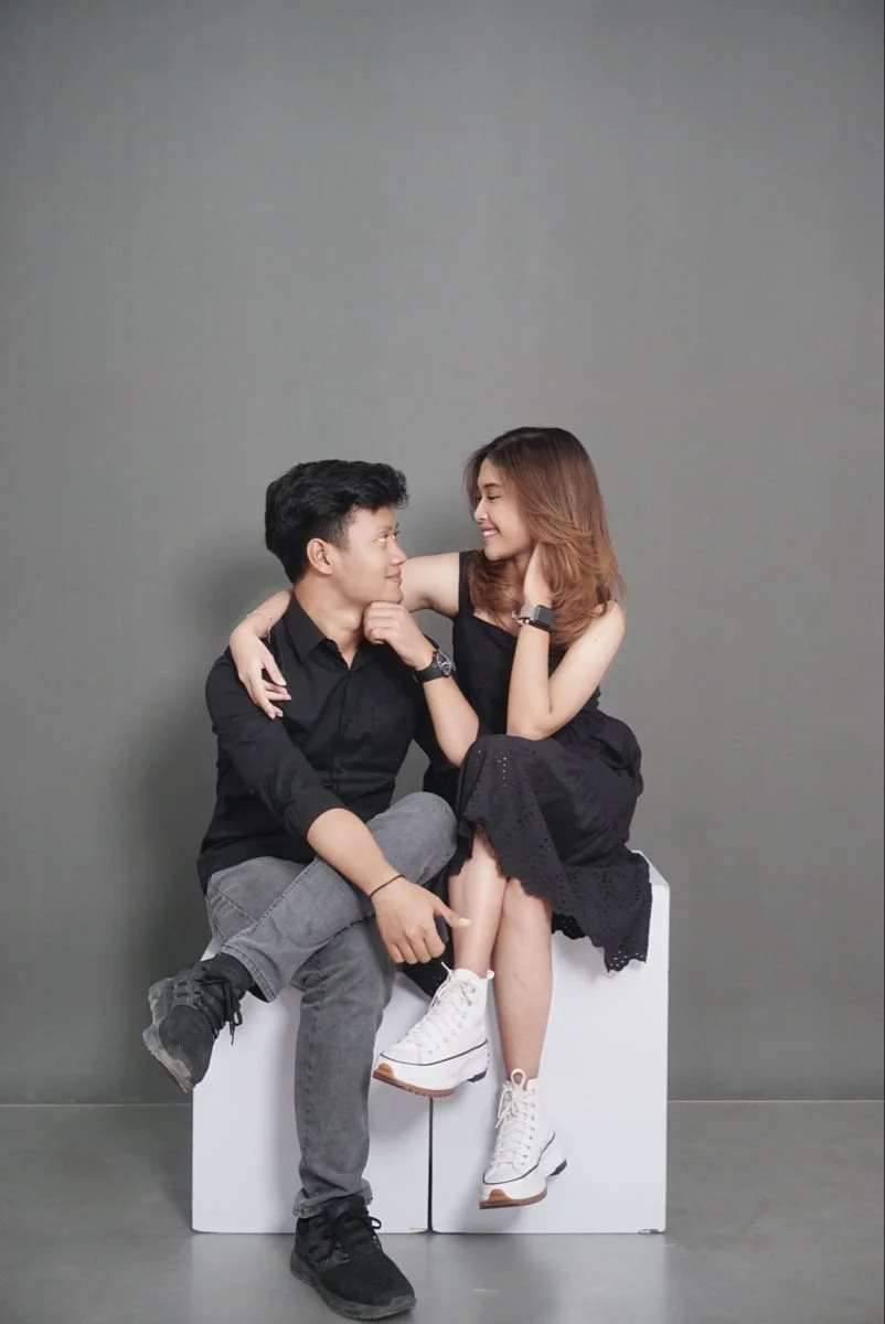 A young man and woman sitting closely together on a white block, gazing into each other's eyes, with the woman smiling and touching the man's shoulder while the man touches his chin, against a plain gray background.