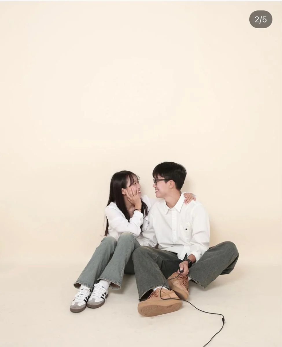A young couple sitting on the floor against a plain, light-colored wall, smiling and looking at each other. The woman has her hand on the man's face, and he has his arm around her shoulder. They are casually dressed, with the woman wearing gray jeans and sneakers, and the man in gray pants, a white shirt, and brown shoes, holding a wired device.