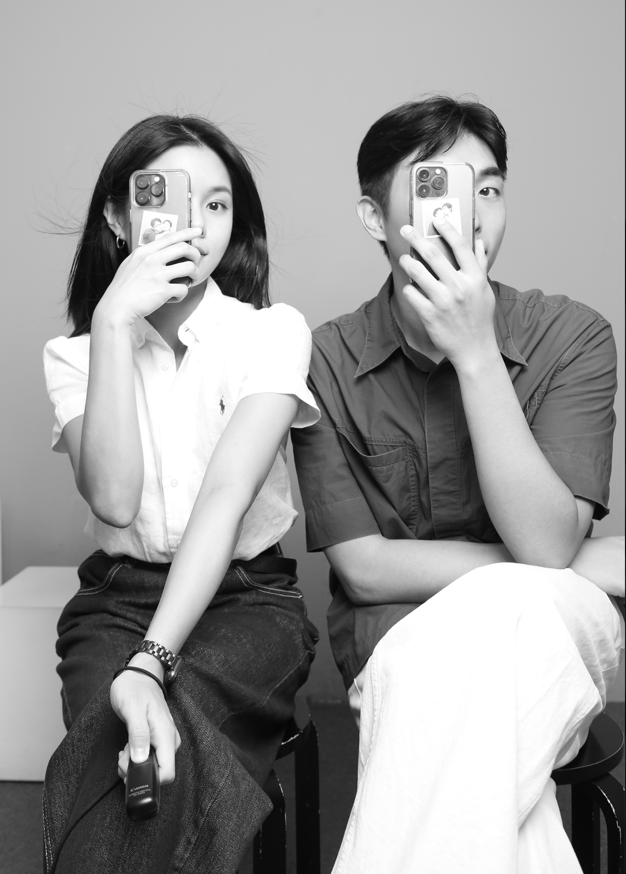 Black and white photo of a woman and man sitting side by side, each holding a smartphone in front of their faces, with a small photo on the phone case.