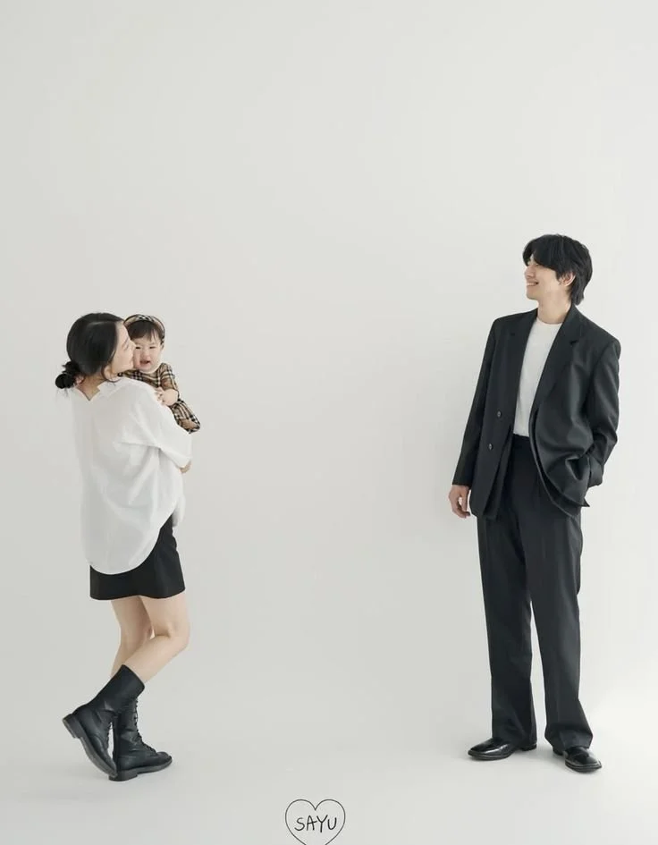 A woman holding a smiling baby in front of a smiling man, all standing against a plain white background.