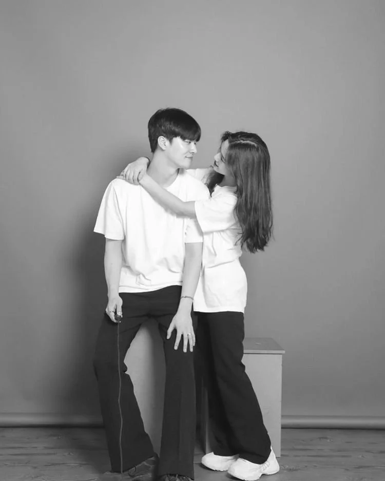 A young man and woman standing close together, facing each other, with the woman embracing the man's shoulders and looking into his eyes against a plain background.