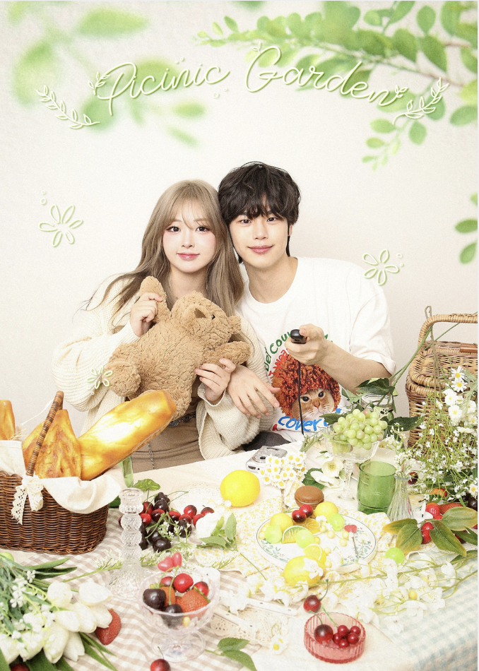 A young woman and man at a picnic table decorated with flowers, fruit, bread, and greenery, holding a teddy bear and a remote control. Behind them, a sign reads 'Picnic Garden'.