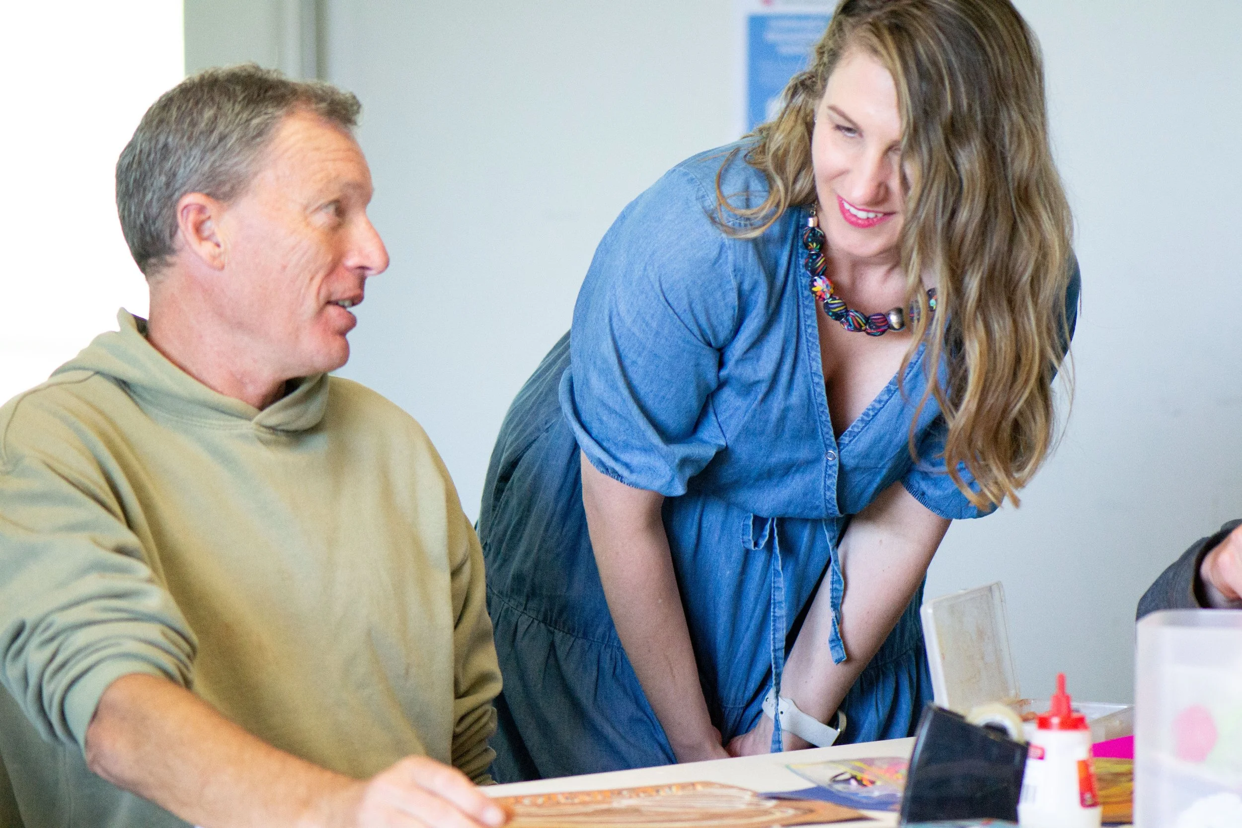 A woman in a blue dress leaning over a man in a beige hoodie, engaged in conversation at a table with arts and crafts supplies.