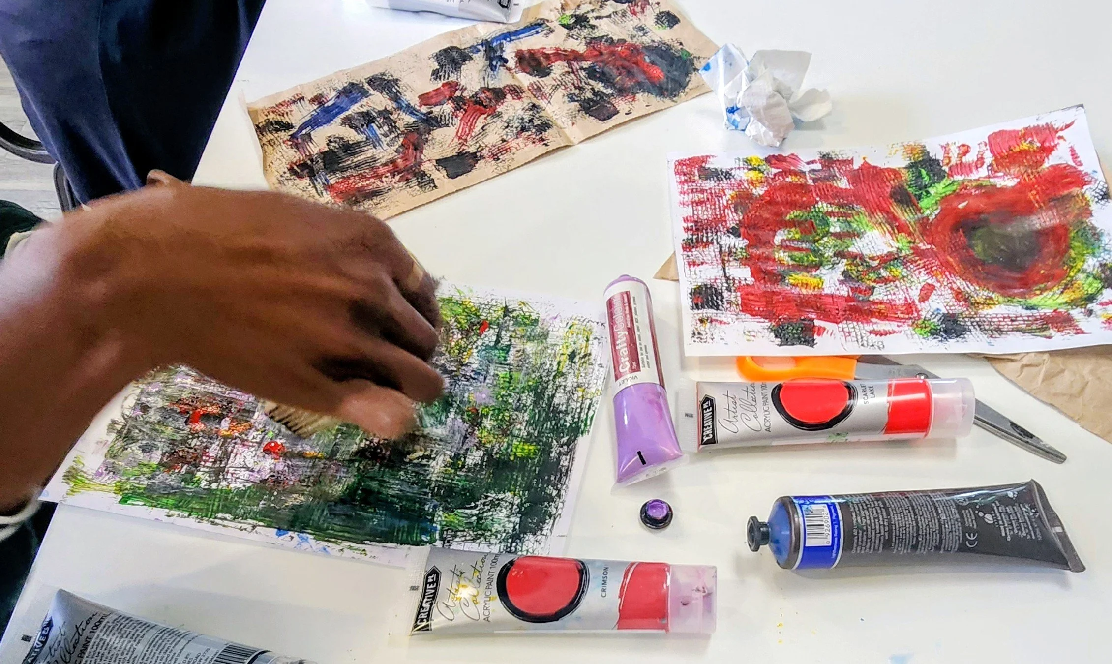 The image shows a person's hand creating abstract artwork with red, black, green, and yellow paint on paper. Various art supplies such as acrylic paint tubes, a paint roller, and brushes are scattered on the white table. There are also crumpled tissues and additional paintings or sketches on the table.