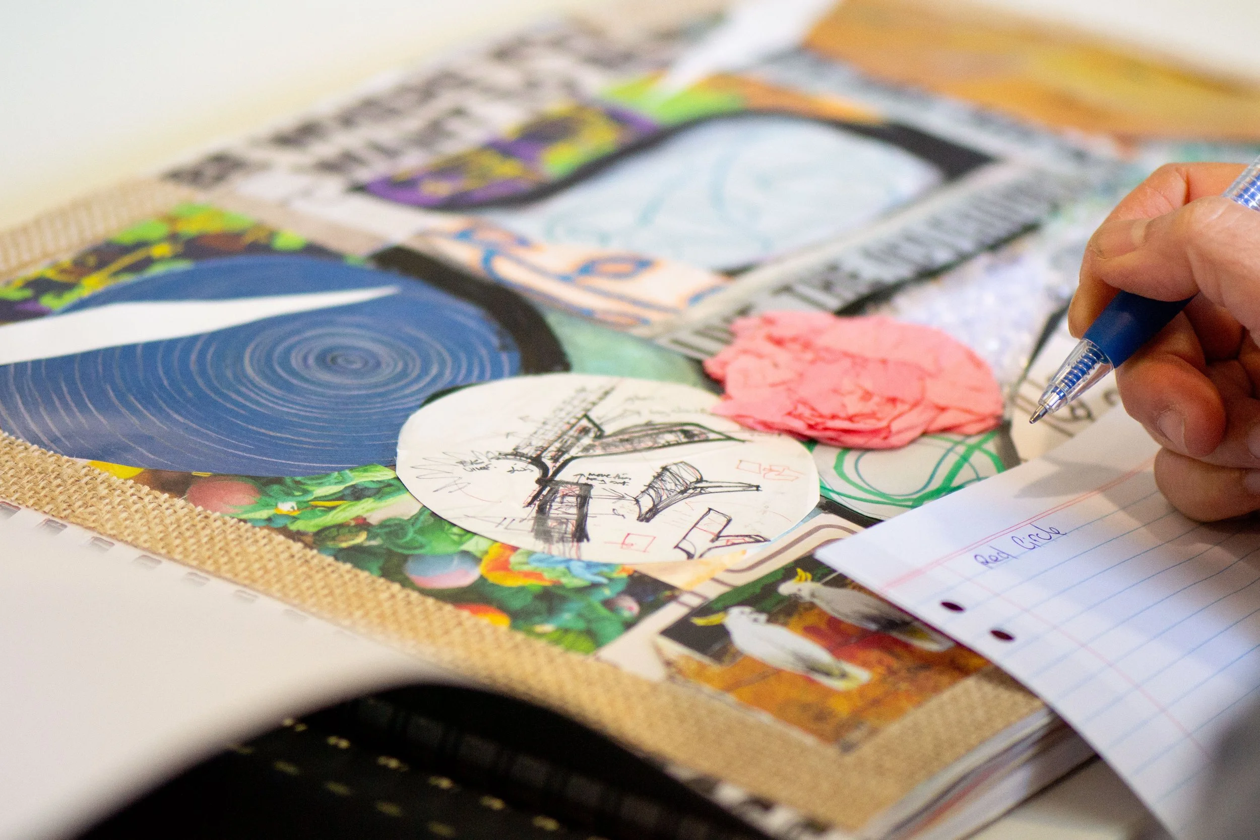 Person sketching and taking notes on a notebook page with sketches and colorful papers on the table.