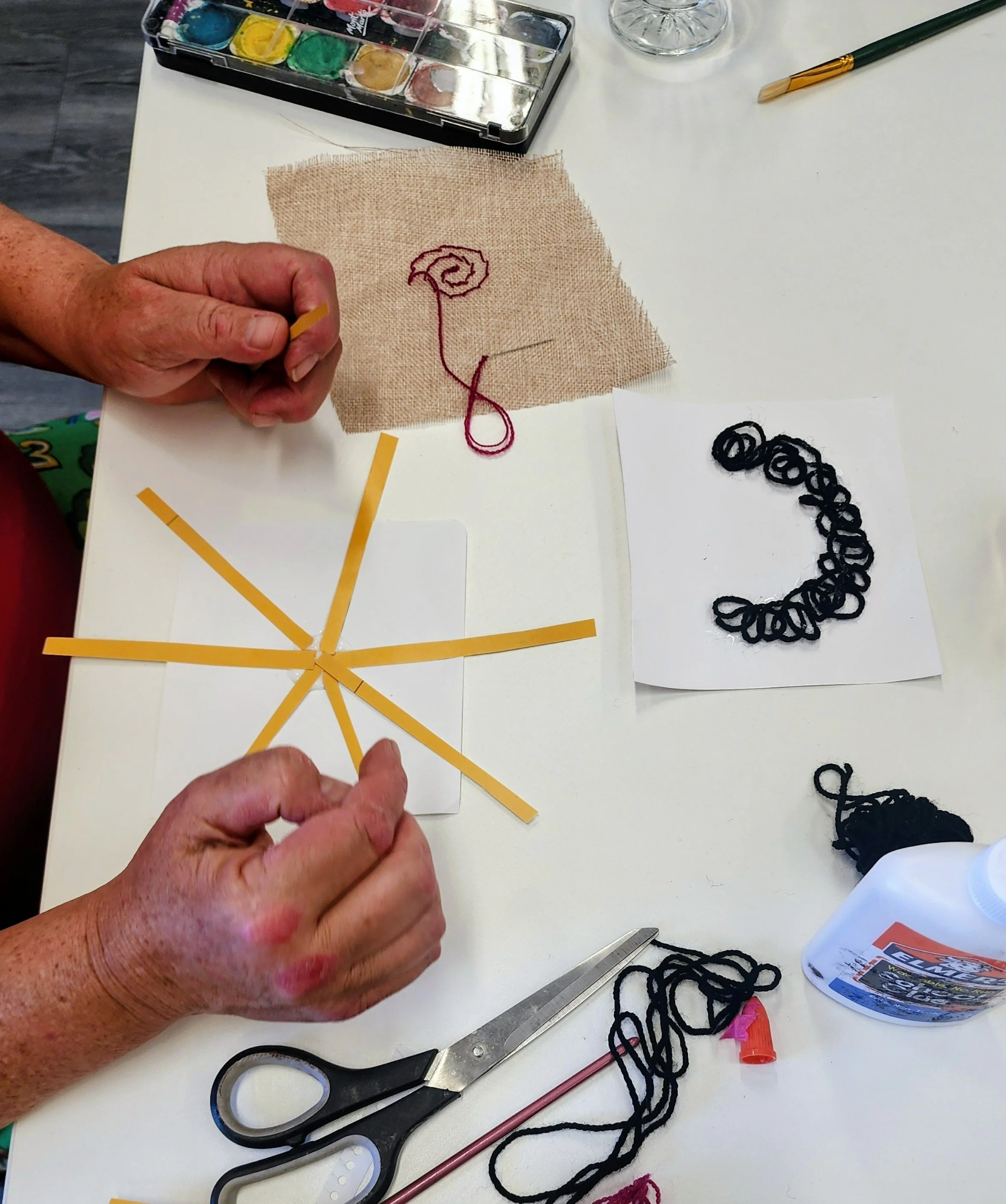 Person creating a craft with yellow paper strips arranged in a star pattern on white paper, black yarn, and embroidery activities on a white table.