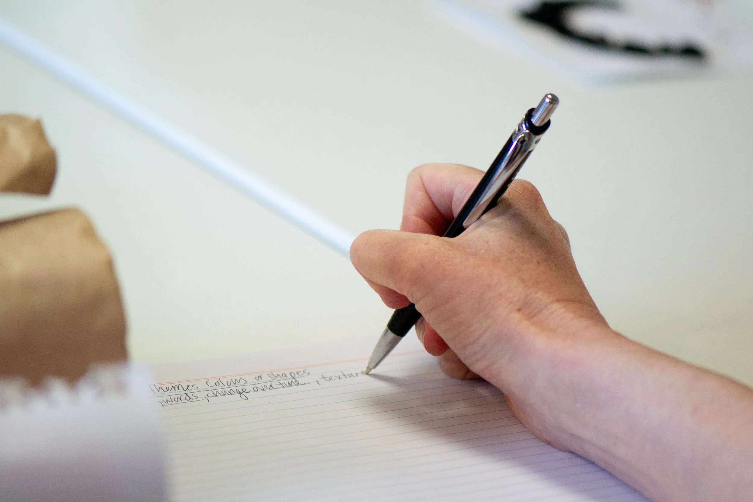 A person's hand holding a silver and black pen writing on lined paper, with a white surface in the background.