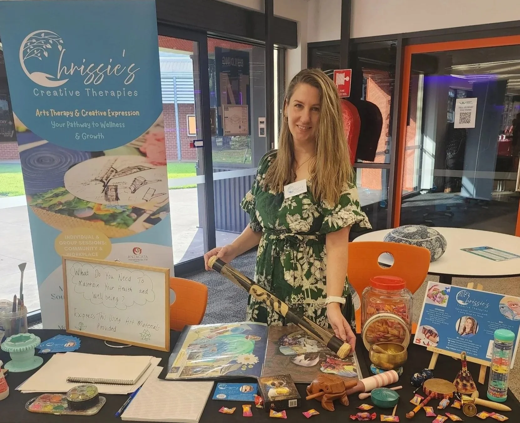 A woman with long blonde hair standing at an arts therapy booth, holding a didgeridoo, with various art supplies and informational materials on display, and a sign reading 'Chrigg's Creative Therapies'.