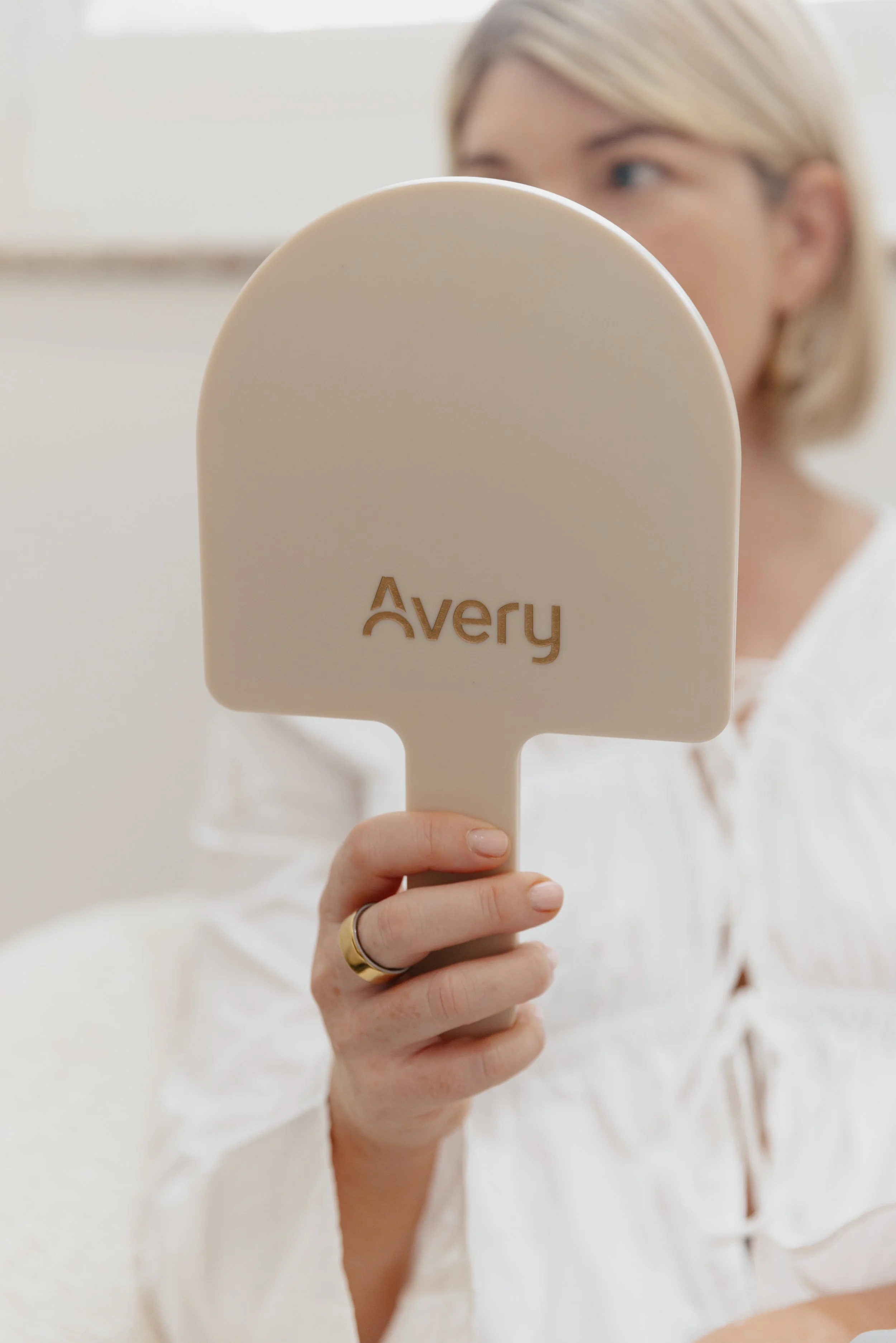 A woman in a white blouse holds an ivory-colored hand mirror with the brand name 'Avery' on the back, partially covering her face.