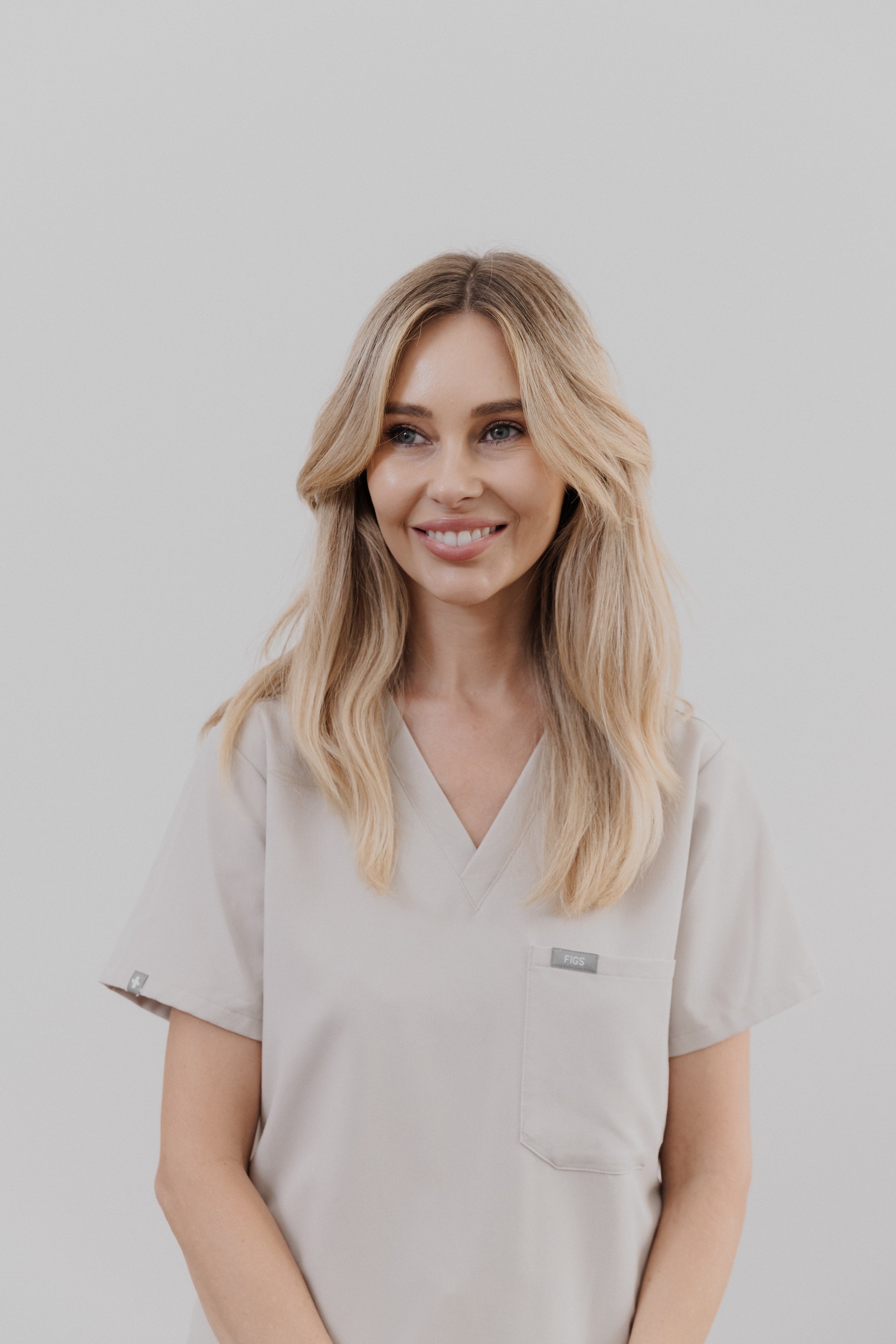A woman with blonde wavy hair smiling, wearing a light beige scrub top with a pocket labeled "FIGS," against a plain white background.
