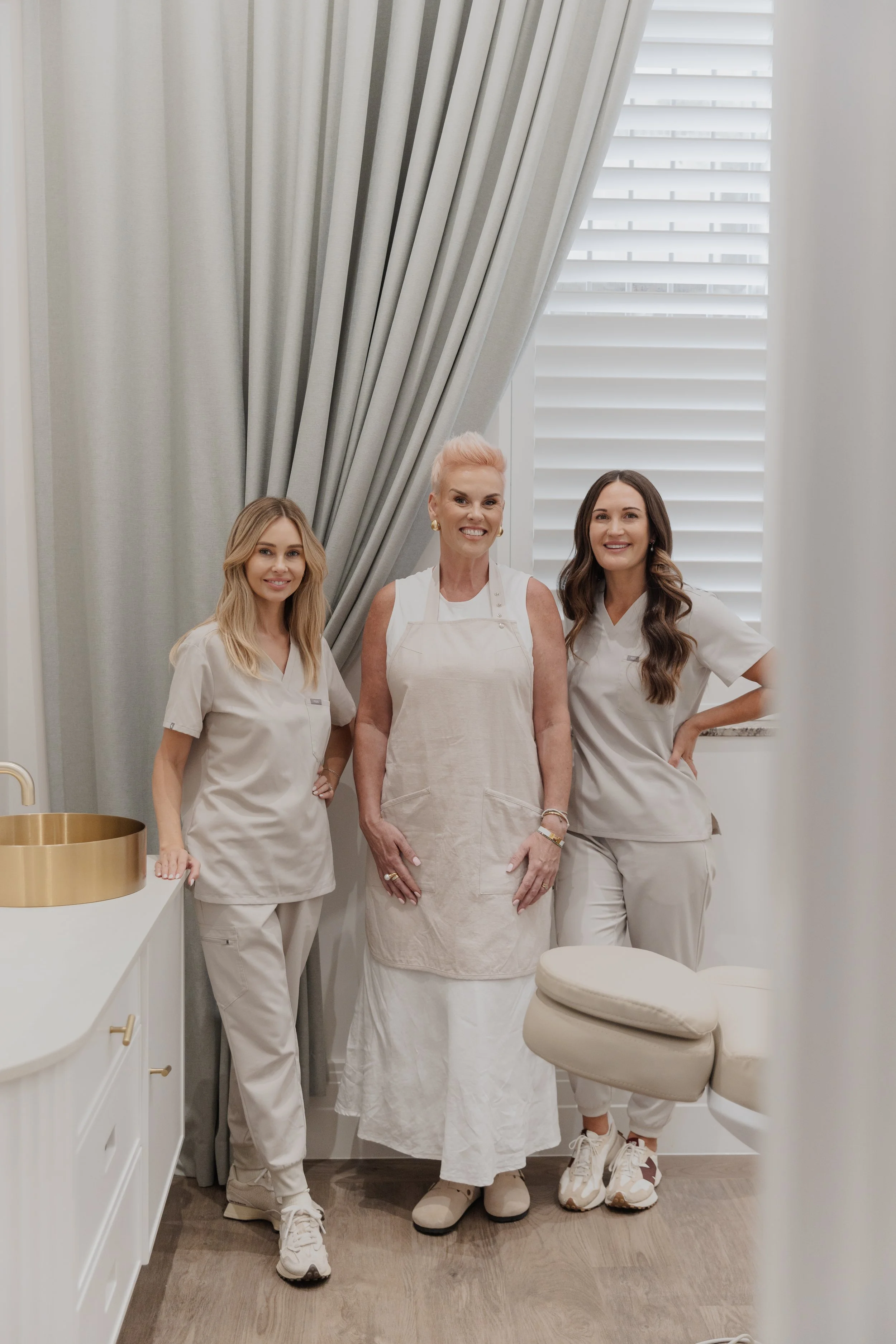 Three women in medical scrubs and apron smiling in a room with white walls, curtains, and window blinds.