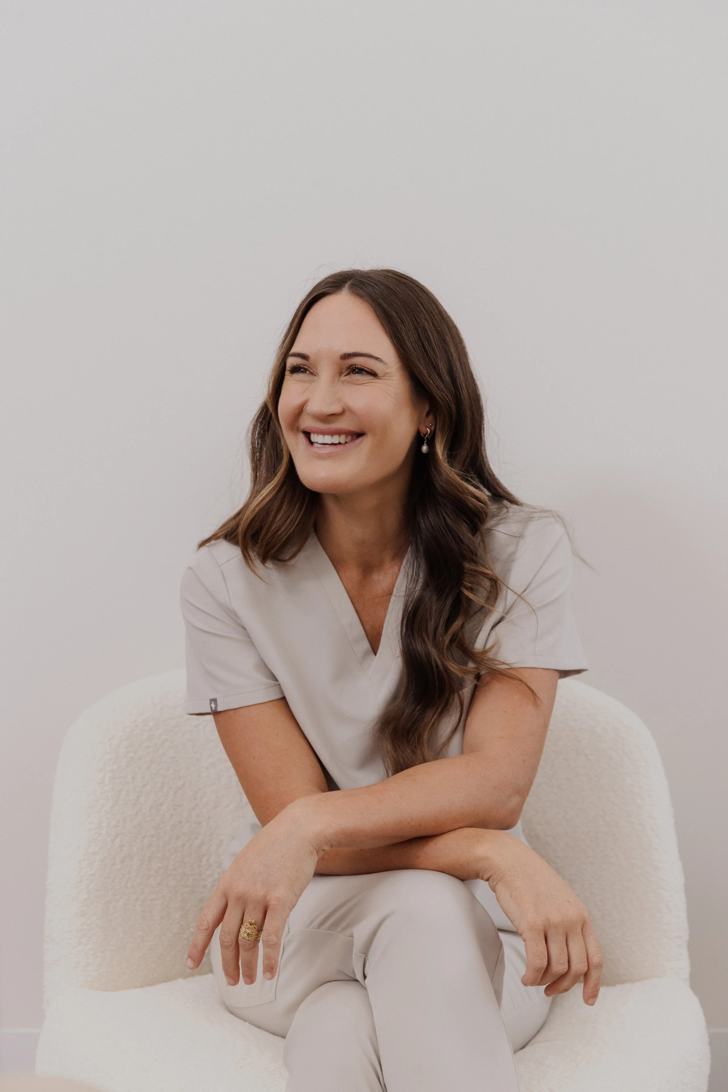 A smiling woman with long brown hair, wearing a light beige medical uniform, sitting on a white textured chair against a plain light-colored background.