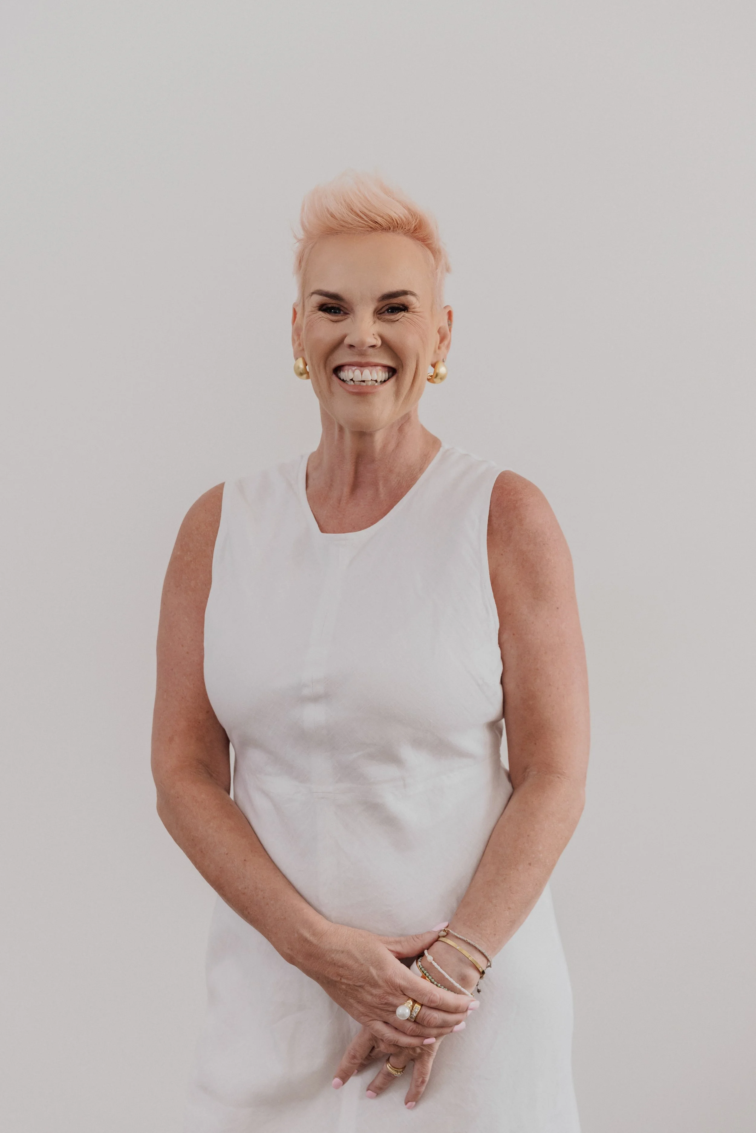A smiling woman with short pink hair, wearing a white sleeveless dress and gold jewelry, standing against a plain white background.