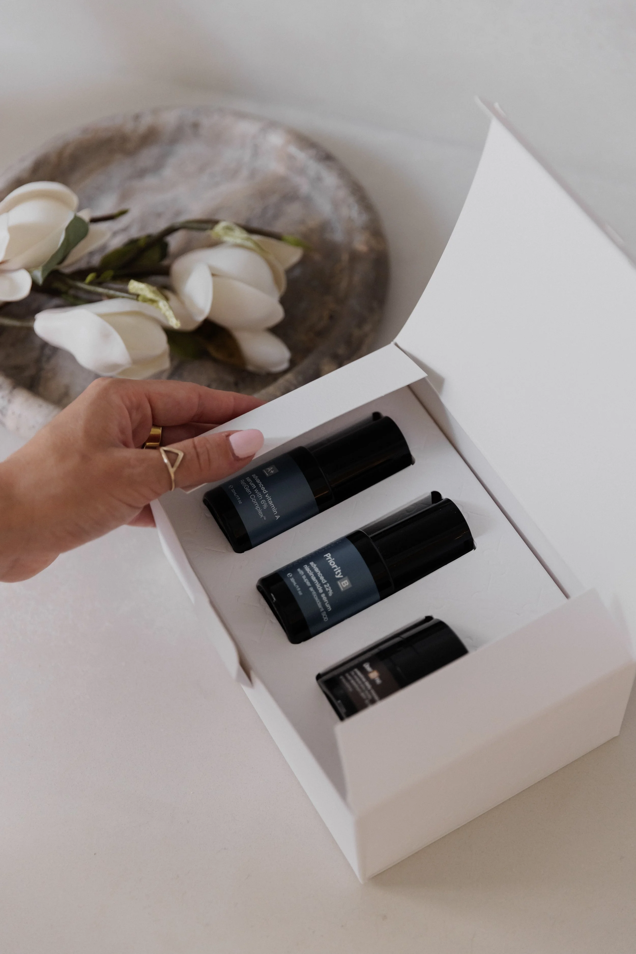 A hand with jewelry opening a white box containing four small black bottles, with a decorative flower arrangement in the background.