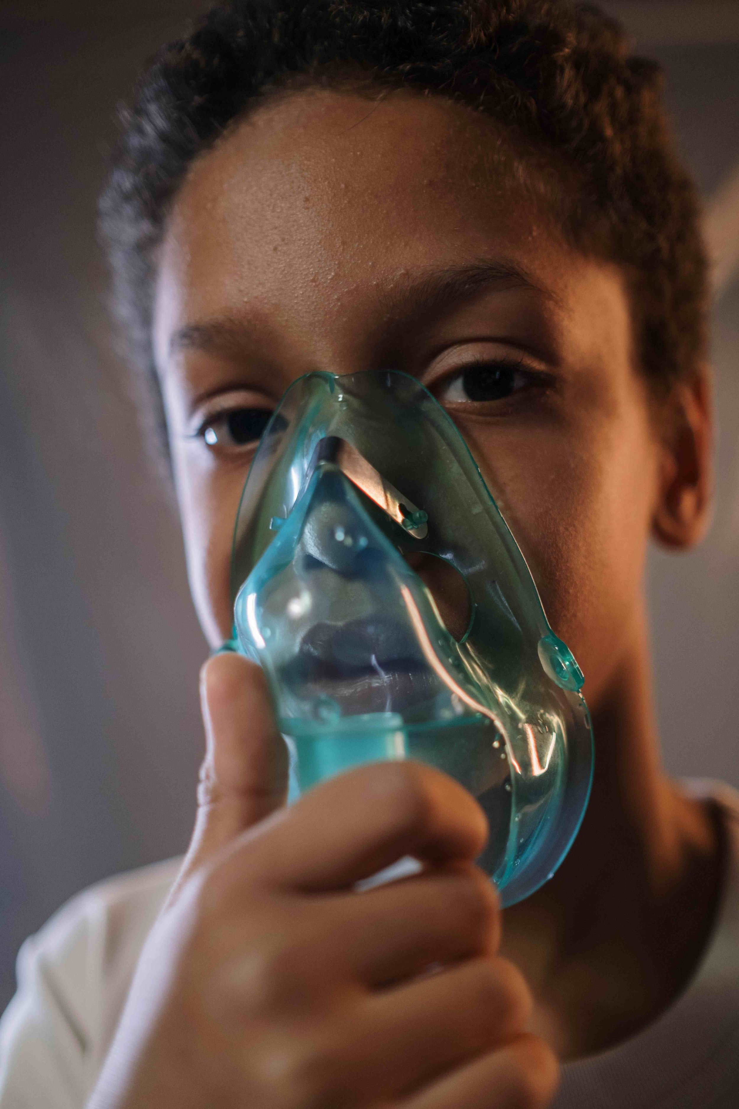 A young person with curly hair wearing a headband holding a turquoise oxygen mask over their face, with their eyes partially closed, in a close-up shot.