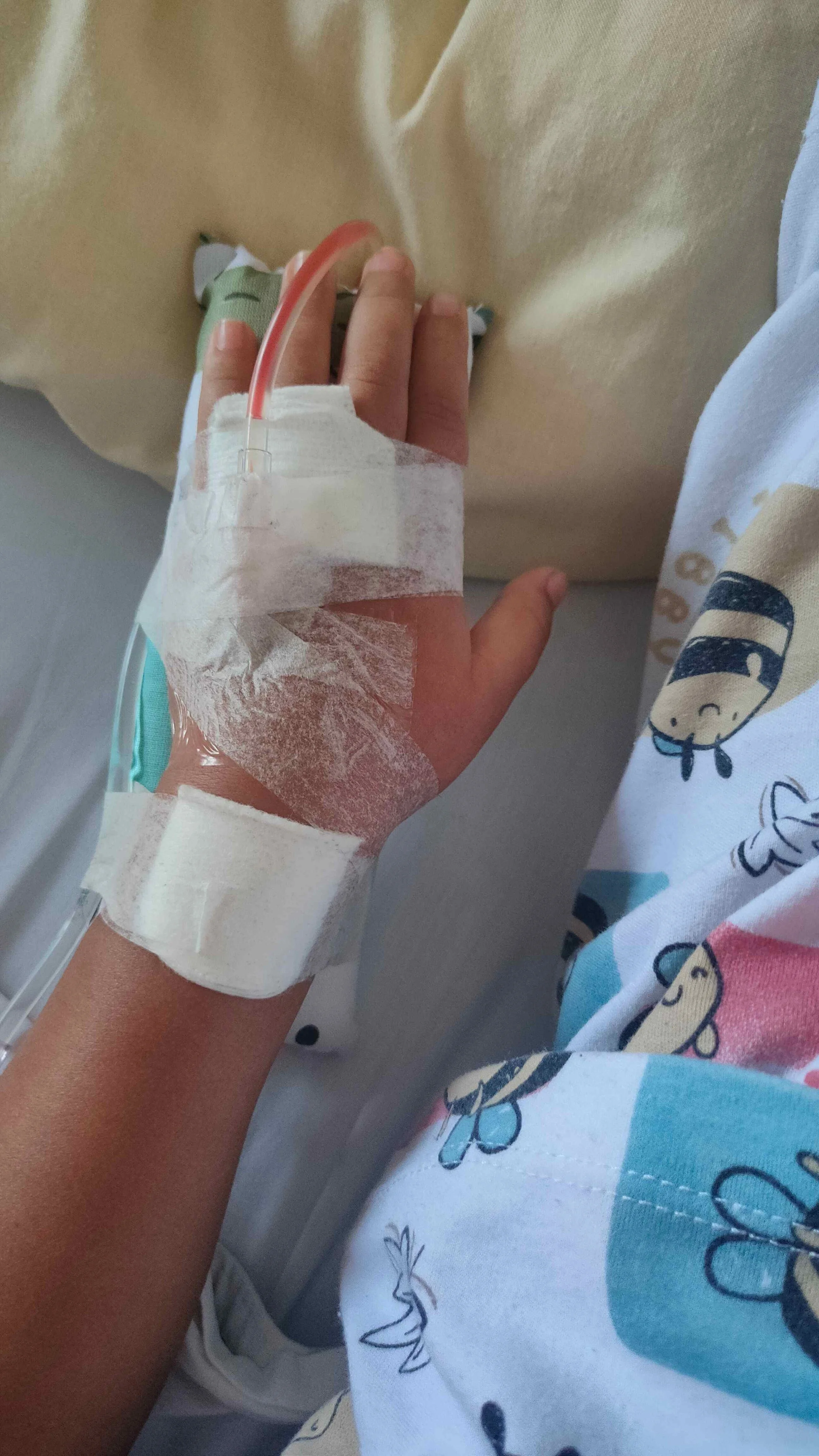 Close-up of a patient's hand with an IV line and bandage, lying in a hospital bed with a colorful cartoon-themed blanket.
